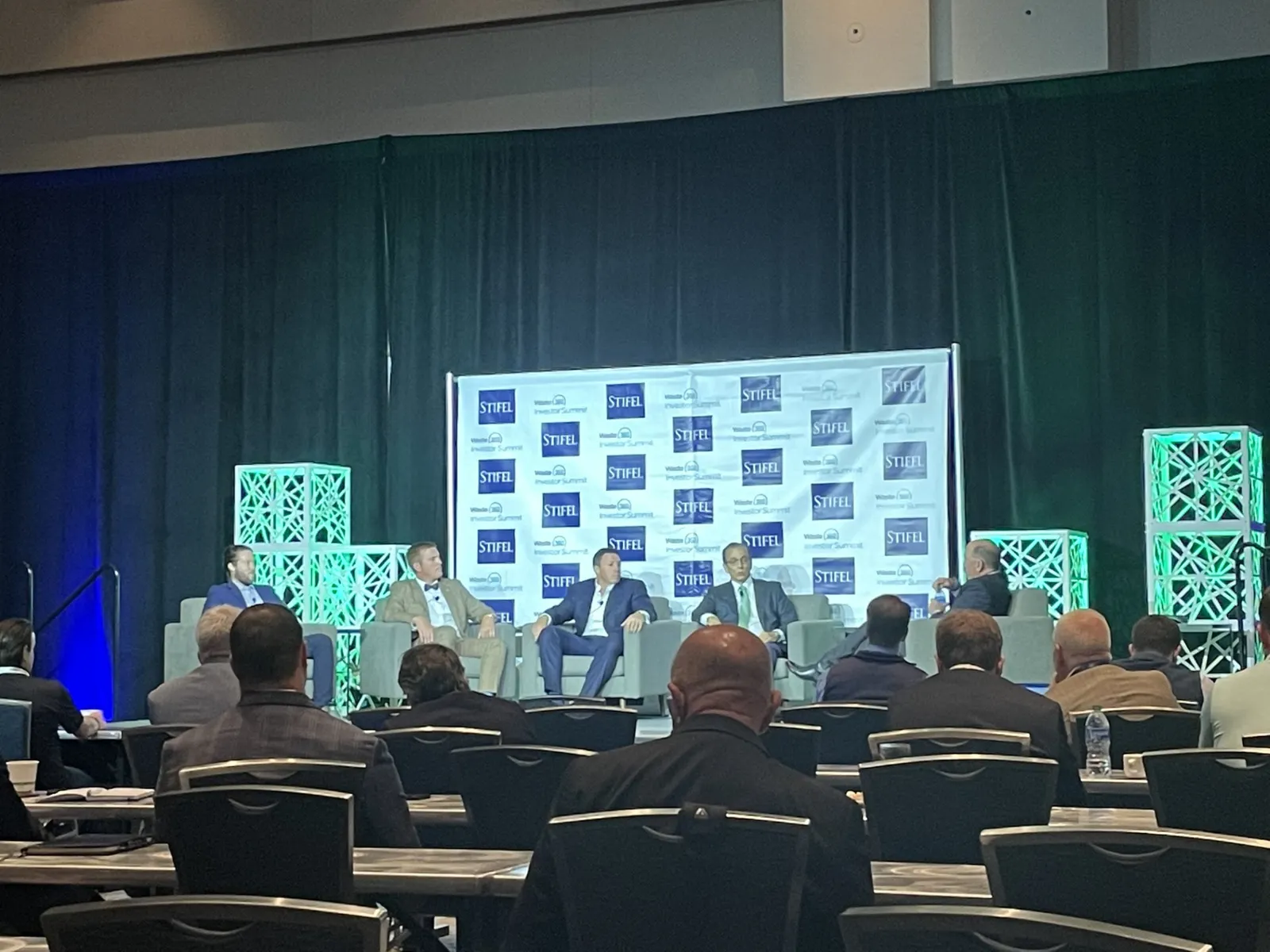 Five people sit in chairs on a stage in front of a room of people in a convention center