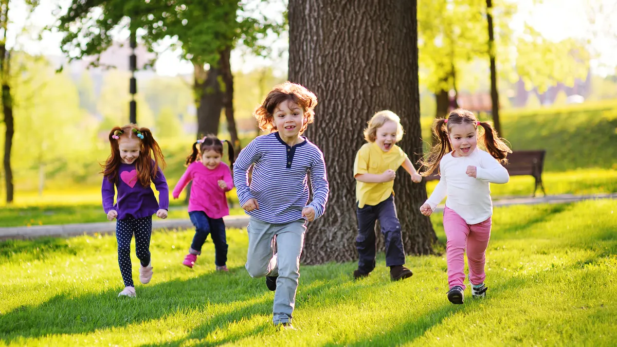 Five young students run toward camera while in a park-like setting with grass, a tree and a bench.