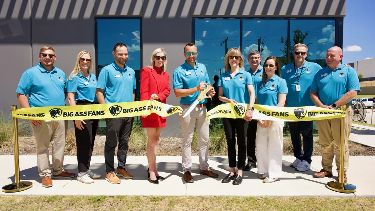 Nine people in blue polos and one person in a red suit behind a yellow ribbon cut in half.