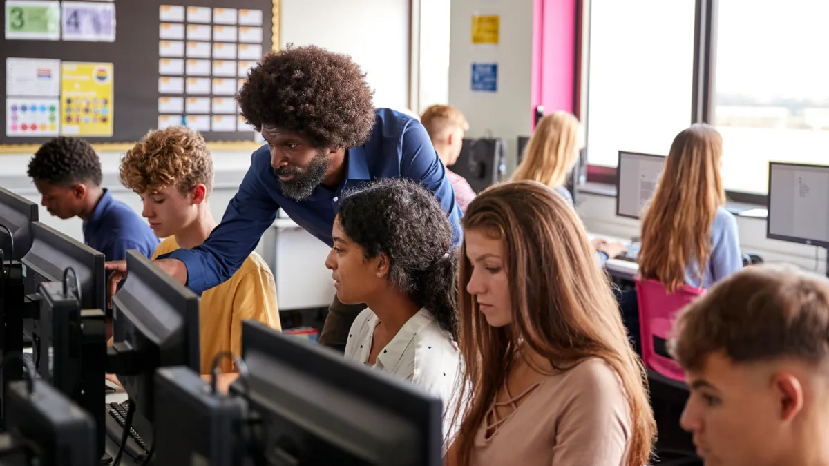 A teacher assists a student working on a computer as their classmates work on nearby computers.