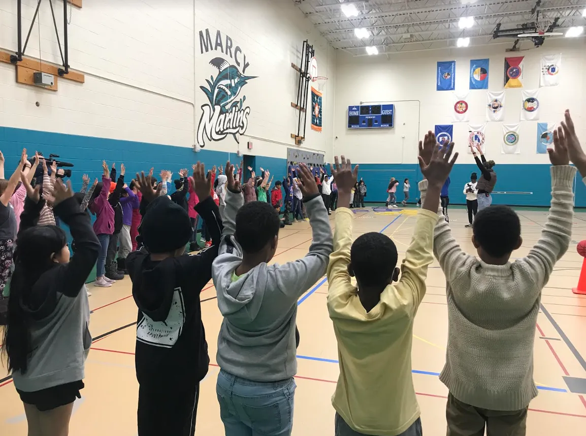 Students stand at the edge of one end of a basketball court in a school gym. Many have their hands raised above their heads.