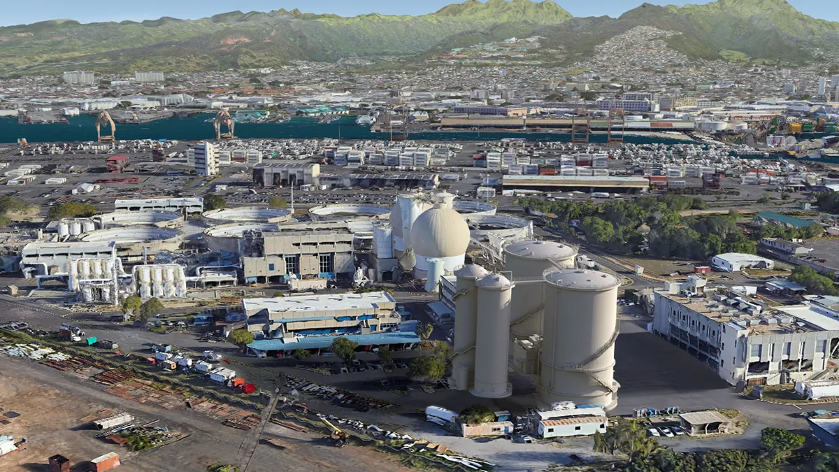 Two towering white cylinders sit among other wastewater treatment buildings, with green mountains in the background.