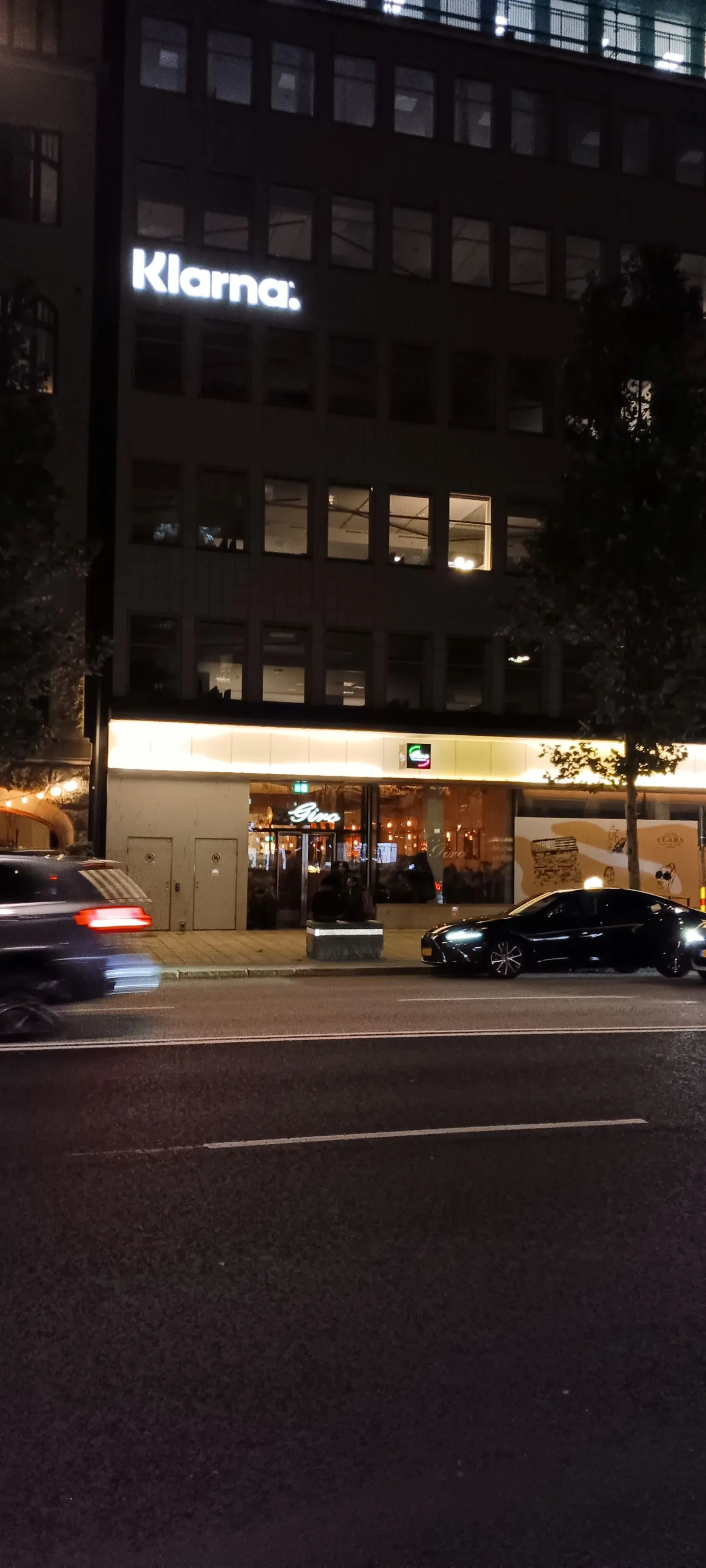 Klarna&#x27;s Stockholm headquarters at night with car in front on street and company&#x27;s name in lights on building.