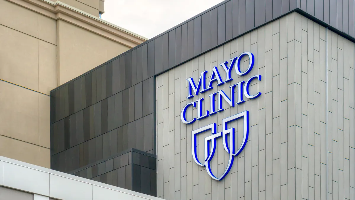 Close-up of a modern building with the Mayo Clinic logo in blue and white, displayed on a vertical tiled wall with part of the adjacent structure visible.