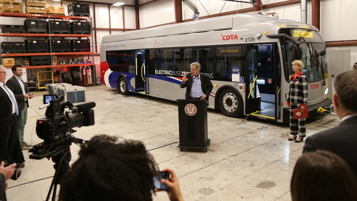 Sen. Sherrod Brown, a gray-haired man wearing a dark suit and blue shirt, stands at a podium in a large garage in front of an electric bus, with others nearby.