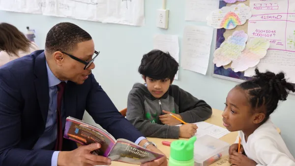 An adult is sitting at a table and holding a book. Two young students are also seated at the desk and are looking at the book and holding pencils.