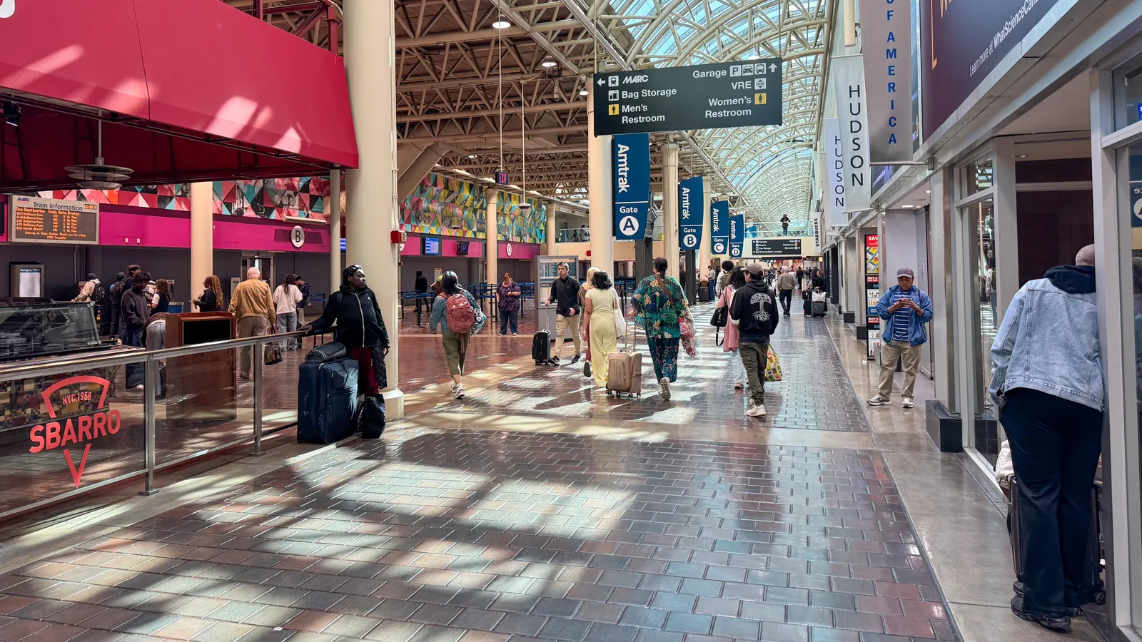 People walking and standing amid a long concourse with shops on the right and train platform access on the left.