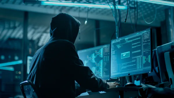 Hooded person types on computer in a dark room with multiple monitors and cables everywhere.