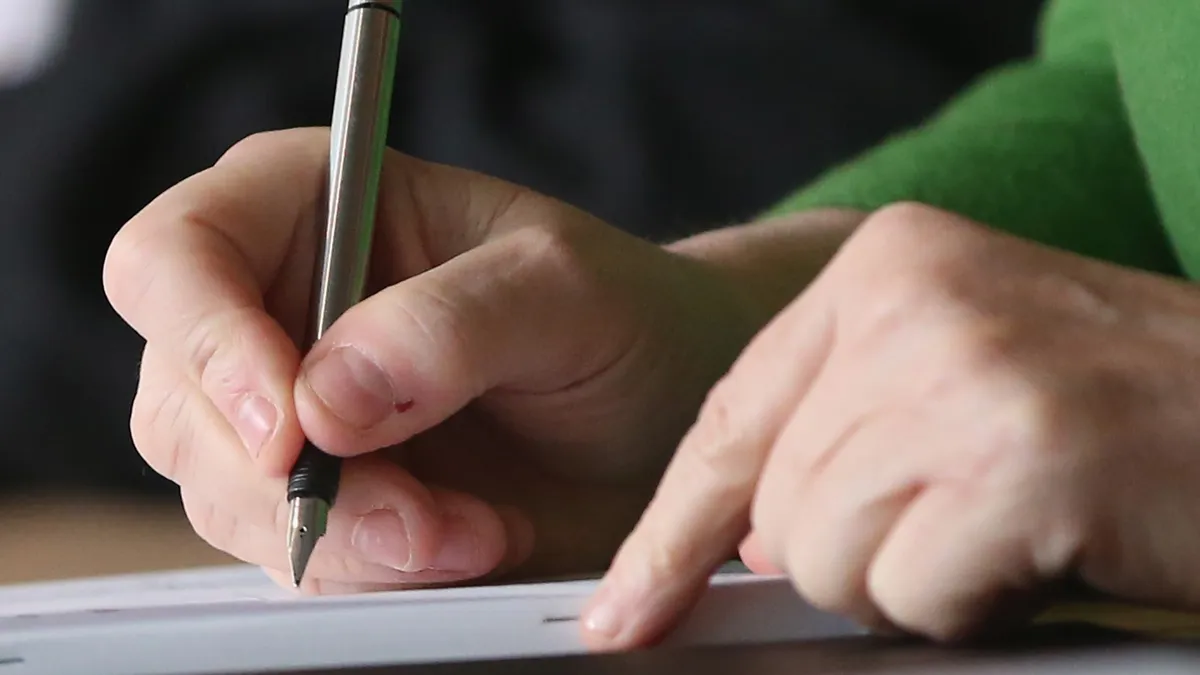 A hand is depicted writing on a piece of paper with a pen.