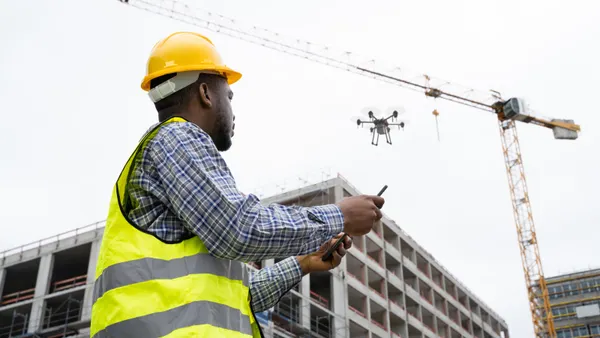 A person in a collared shirt and safety equipment looks up at a drone flying above them.