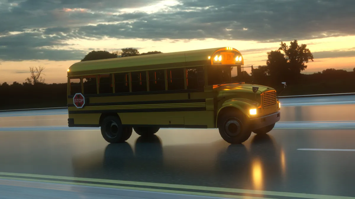 A yellow school bus drives on a road during dusk or dawn with its headlights on, passing a wooded area.