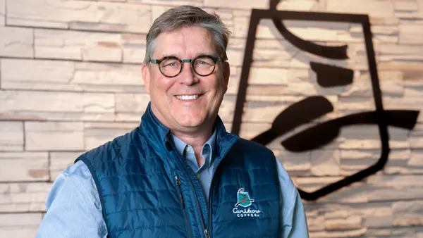 An image of a person holding an iced coffee drink and standing in front of a beige brick building with a Caribou Coffee logo.
