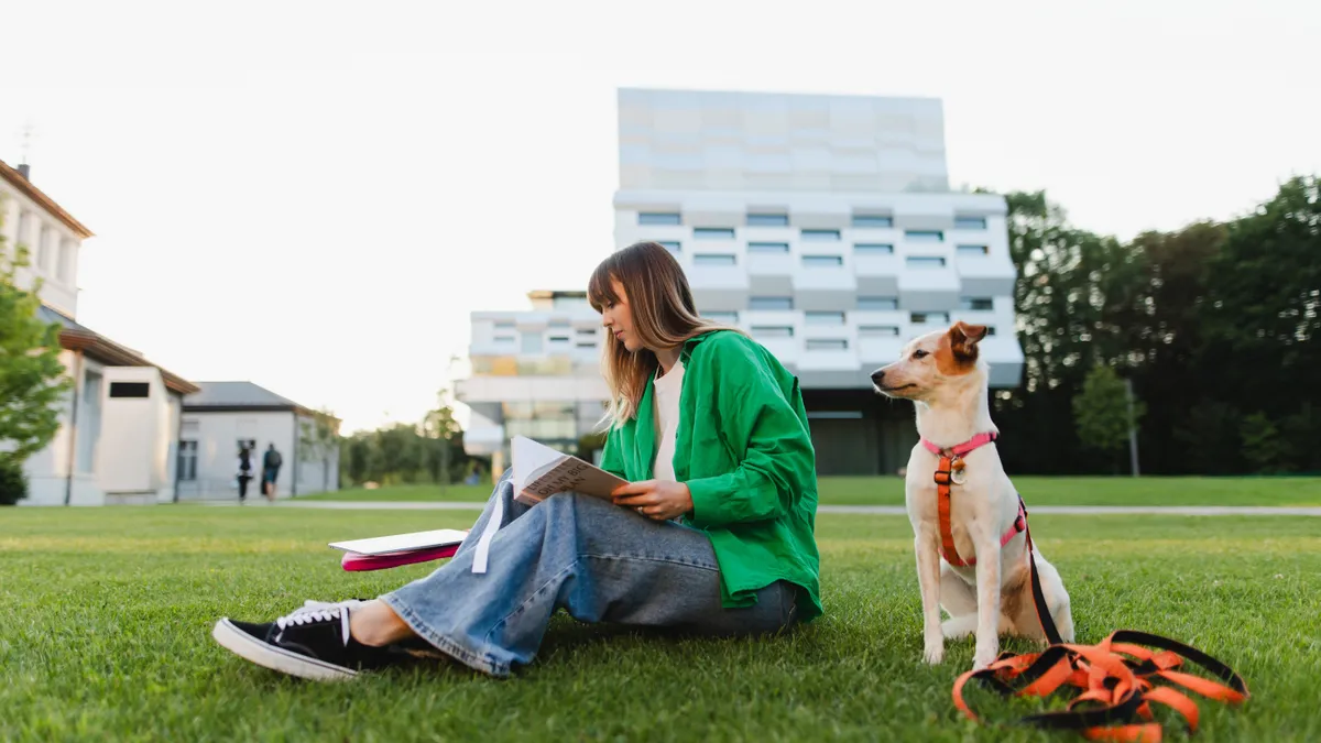 A college student reads a book on the campus lawn while a dog sits nearby.