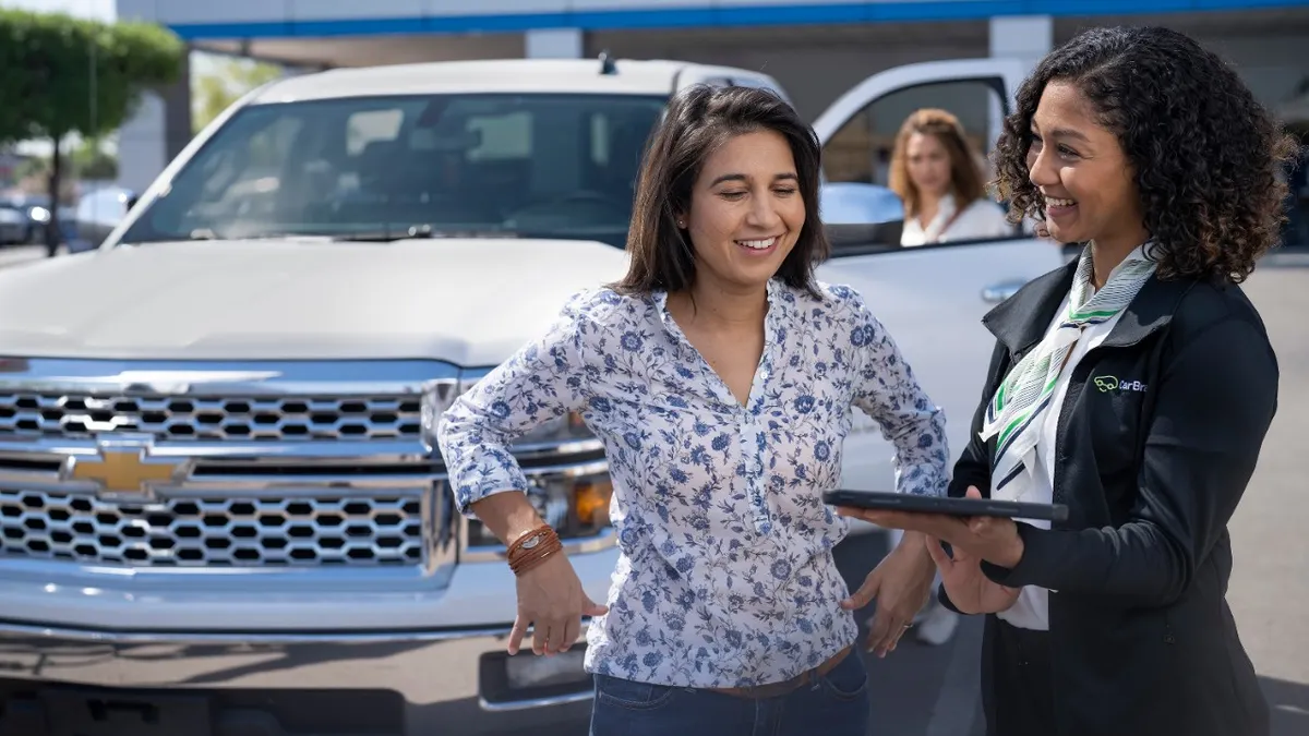 A CarBravo salesperson smiles as she speaks with a potential customer at a GM dealer with a Chevrolet pickup truck in the background.