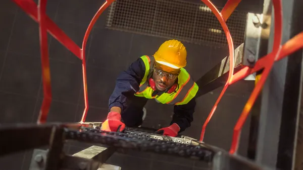 A worker climbing an industrial emergency ladder, wearing a yellow hard hat and reflective vest, and safety glasses.