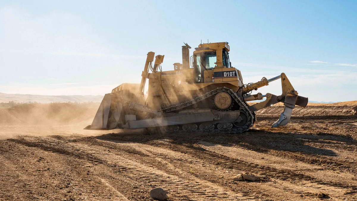 A bulldozer with no pilot on top of hard, dusty ground on a sunny day. There is dust kicked up by the machinery.