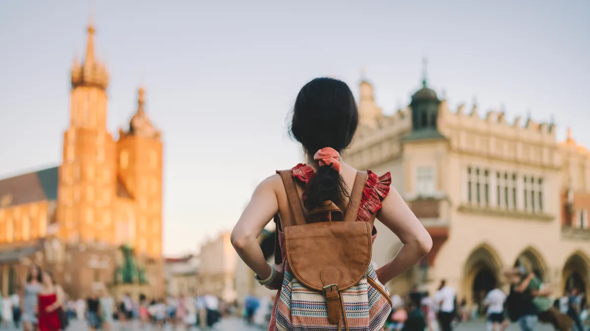 A woman with long dark hair wearing a backpack faces away from the camera, toward a Polish cityscape.