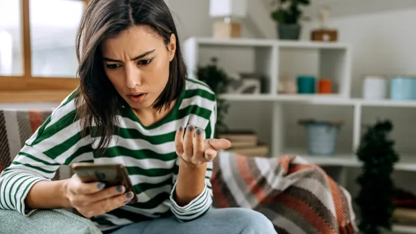 Frustrated young woman with a smartphone in her hands while she sits on a couch at home.