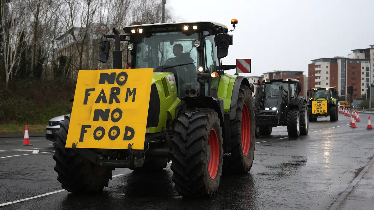 A tractor with a sign that says "No Farm, No Food" leads a caravan of protesters down a street in Wales.