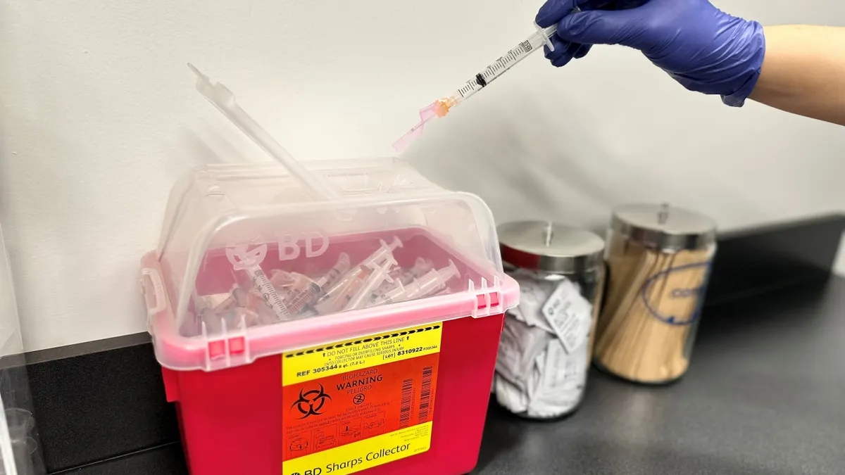 A person wearing a blue glove puts a syringe into a red medical waste container.