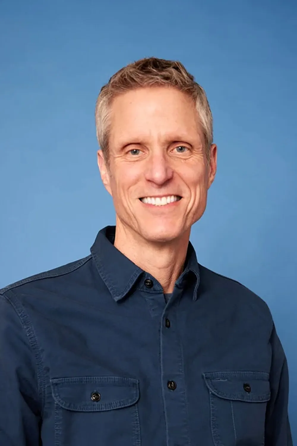 A smiling white man in a blue workshirt against a blue background.