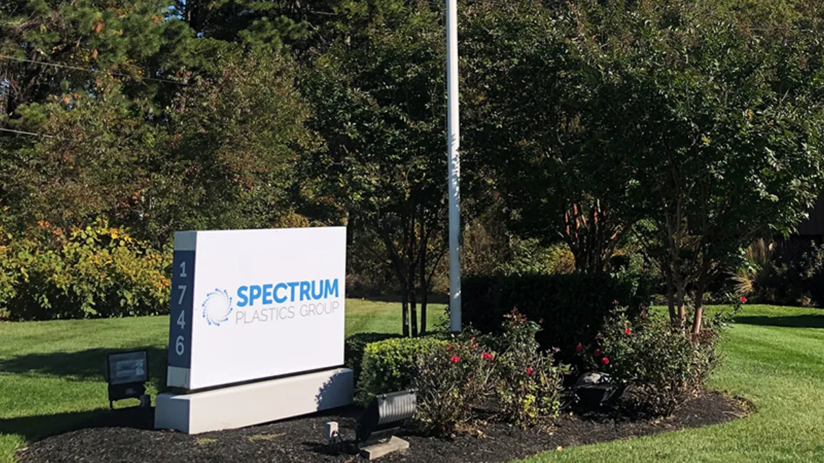 A white sign with Spectrum Plastics Group text on the front, and number 1746 on the right side facing out, surrounded by green landscape on a sunny day.