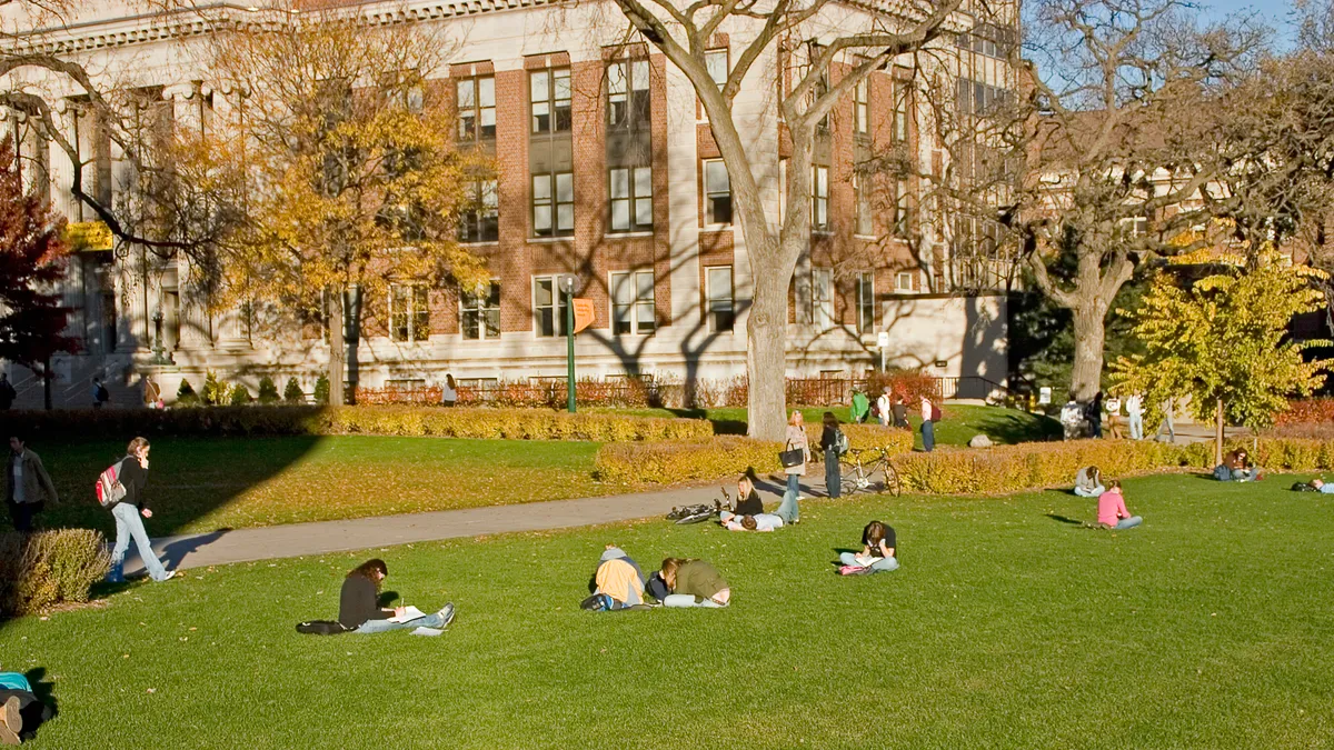 A college quad with students walking and sitting in the grass. Buildings are surrounding the quad.