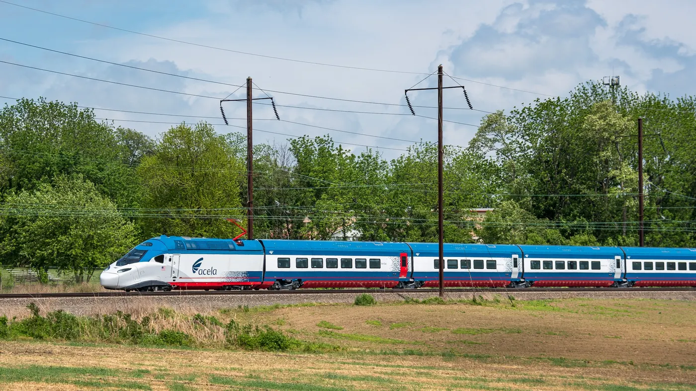 Modern train with blue roof and sides, white band along windows and red stripe along bottom with overhead wires and green trees behind.