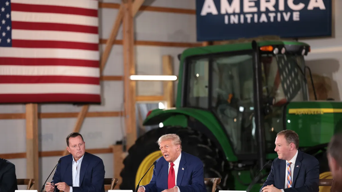 Former President Donald Trump sits at a table next to two men. In the background, a John Deere tractor is seen.