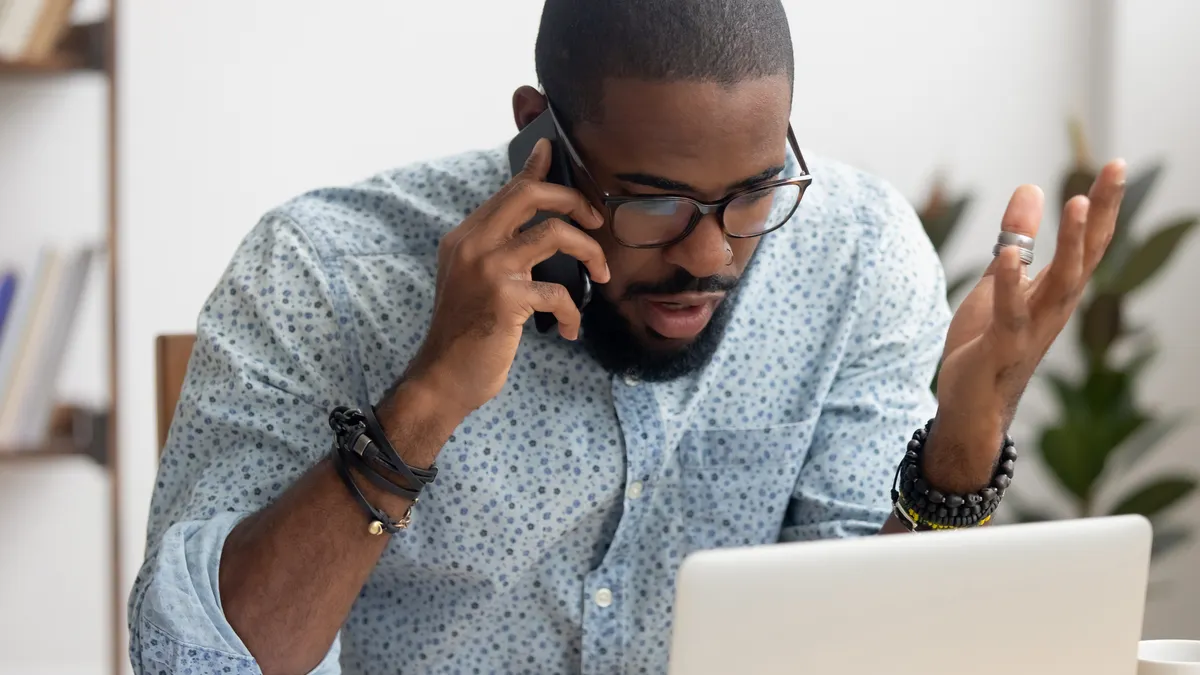 A frustrated man talks on the phone with customer service while looking at his laptop.