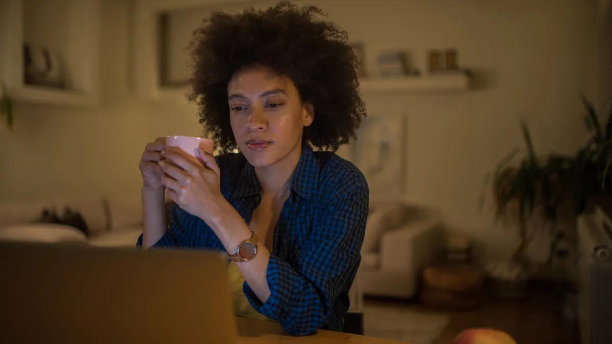 A Black person looks at their laptop, drinking coffee or tea