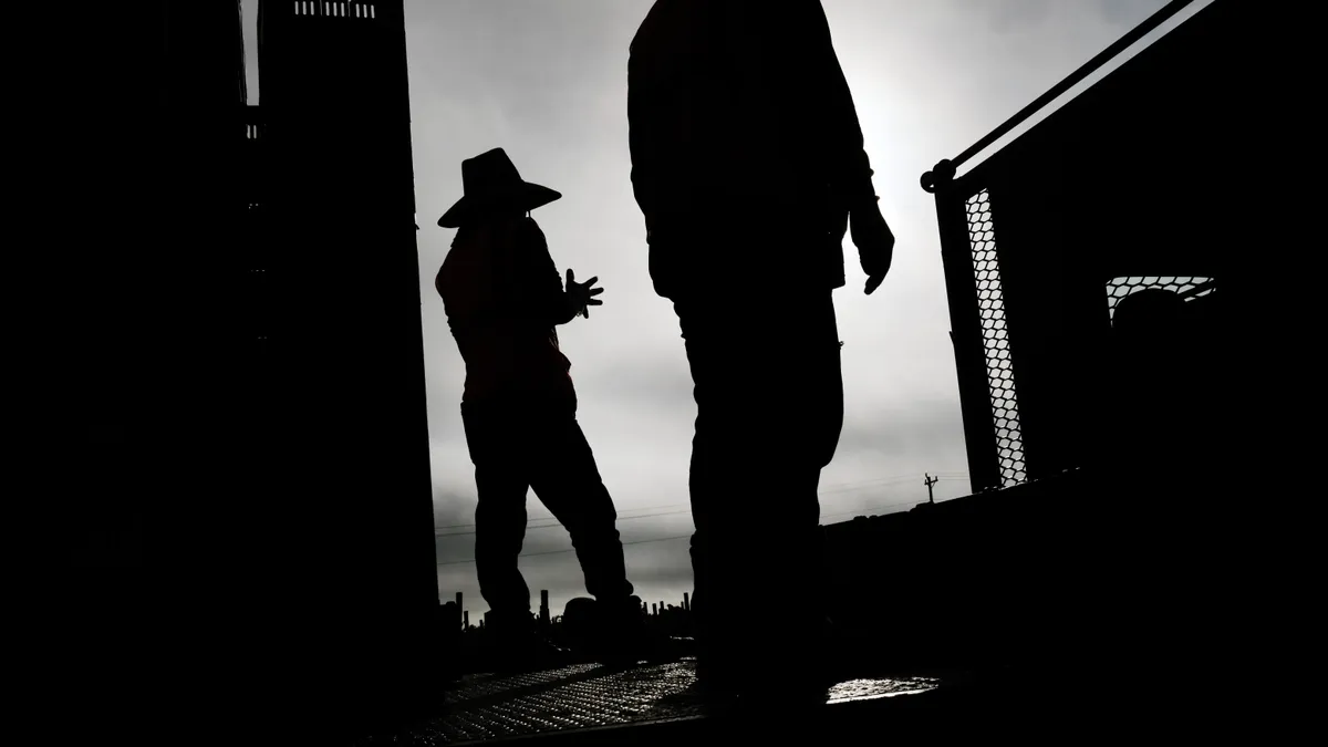 Silhouettes of two men standing in wide-brimmed hats next to a tower of food crates.