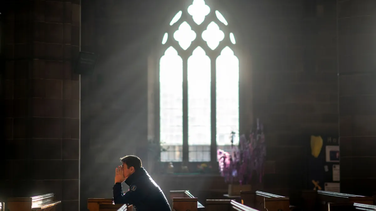 A man in a coat sits in a pew praying while light streams through a window in the background of a stone church.