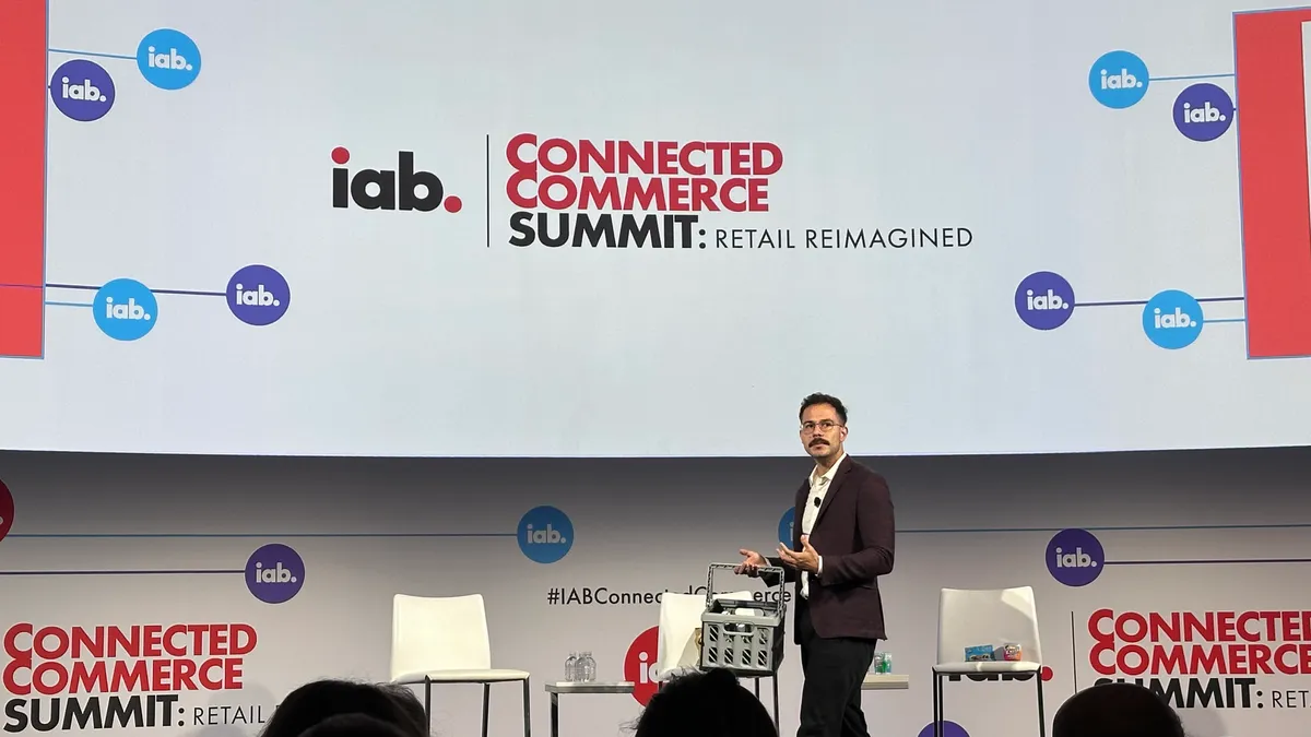 Man on stage with gray grocery basket giving a keynote speak at IAB's Connected Commerce Summit, which is written on a projector screen above