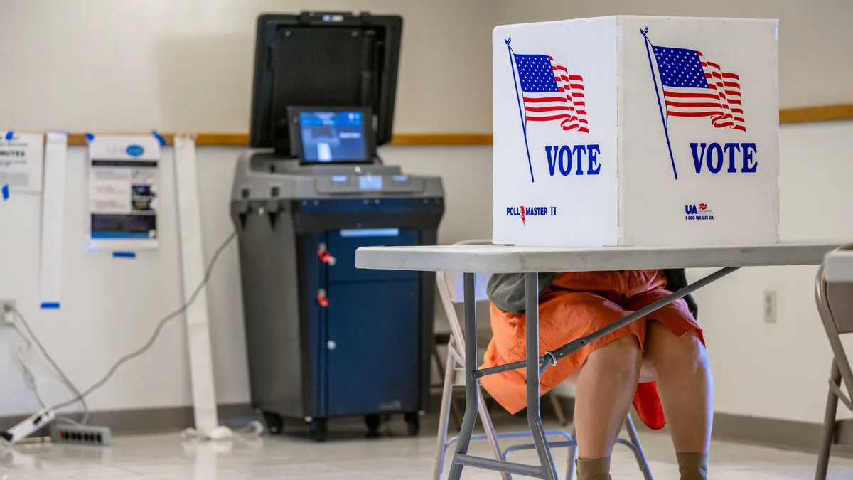 A person sits behind a privacy shield bearing the U.S. flag and the word "vote."