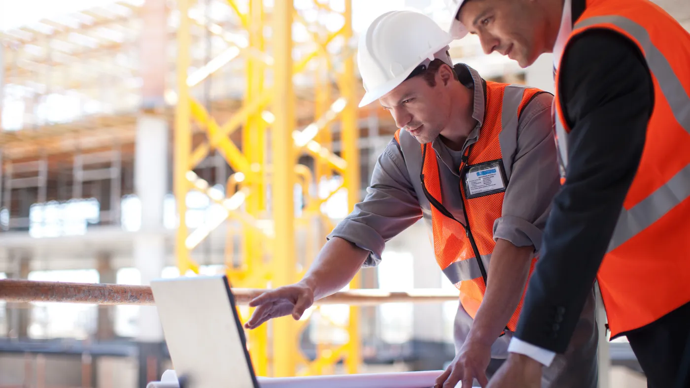 Two people stand in front of a laptop on a construction site while wearing safety gear.