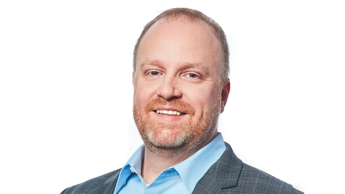 A smiling white man in a blue shirt and a gray jacket with notch lapels.