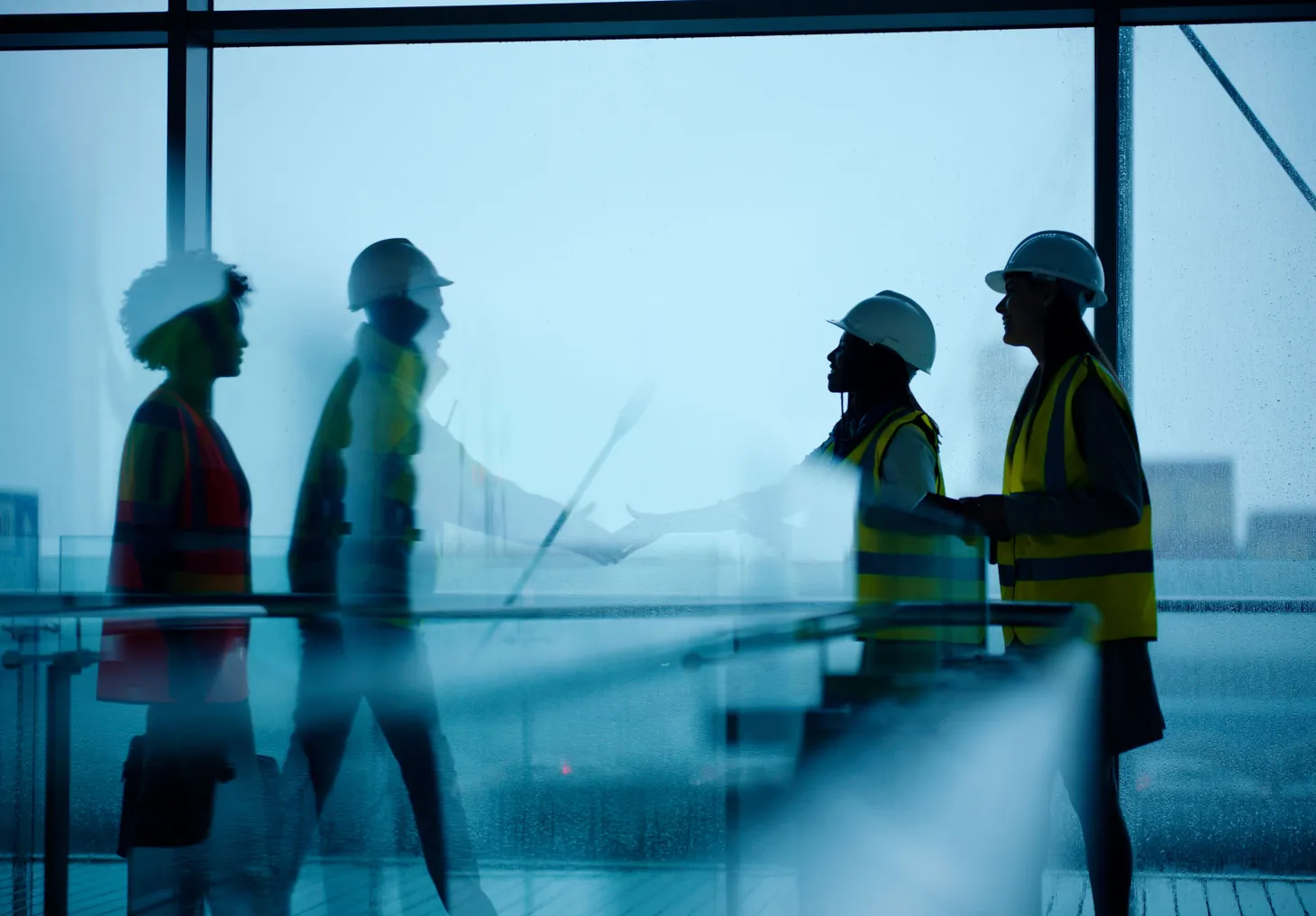 Two pairs of people in construction gear shake hands in a building on a rainy day.