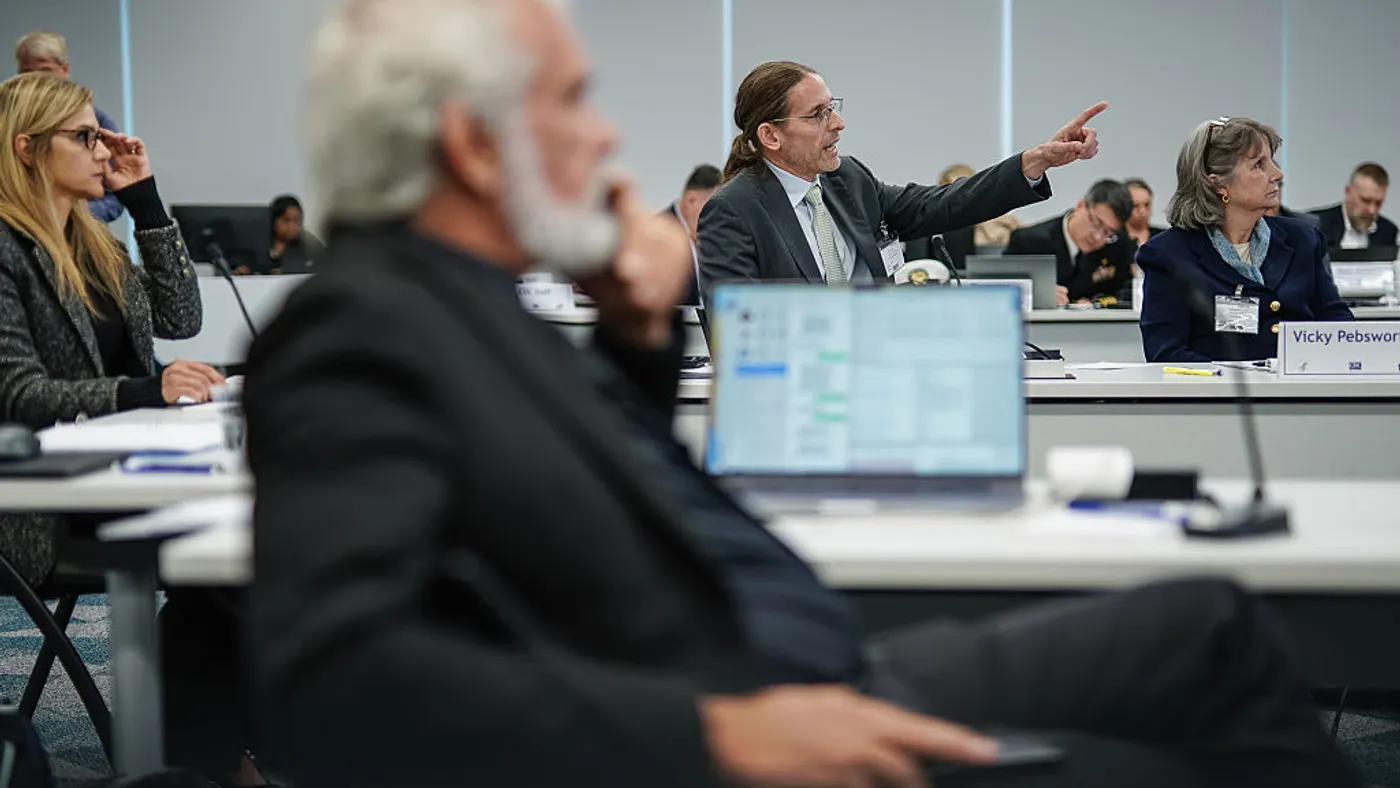 A person in business attire points while seated at a conference table with other people.