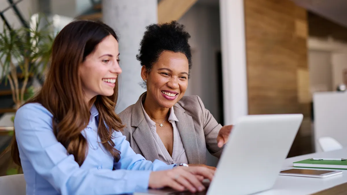 Two businesswomen are collaborating on a project, looking at a laptop and smiling