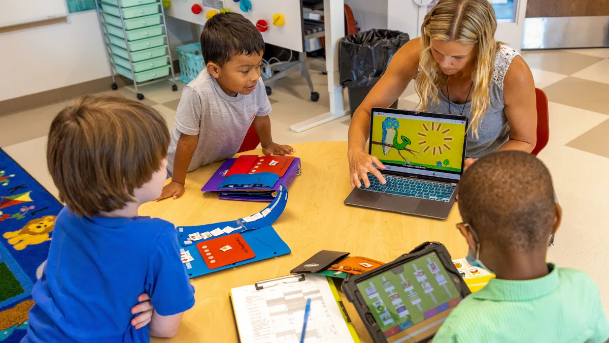 An adult is sitting at a round table in a classroom with young students. The teacher has a laptop open with the screen visible to the students.