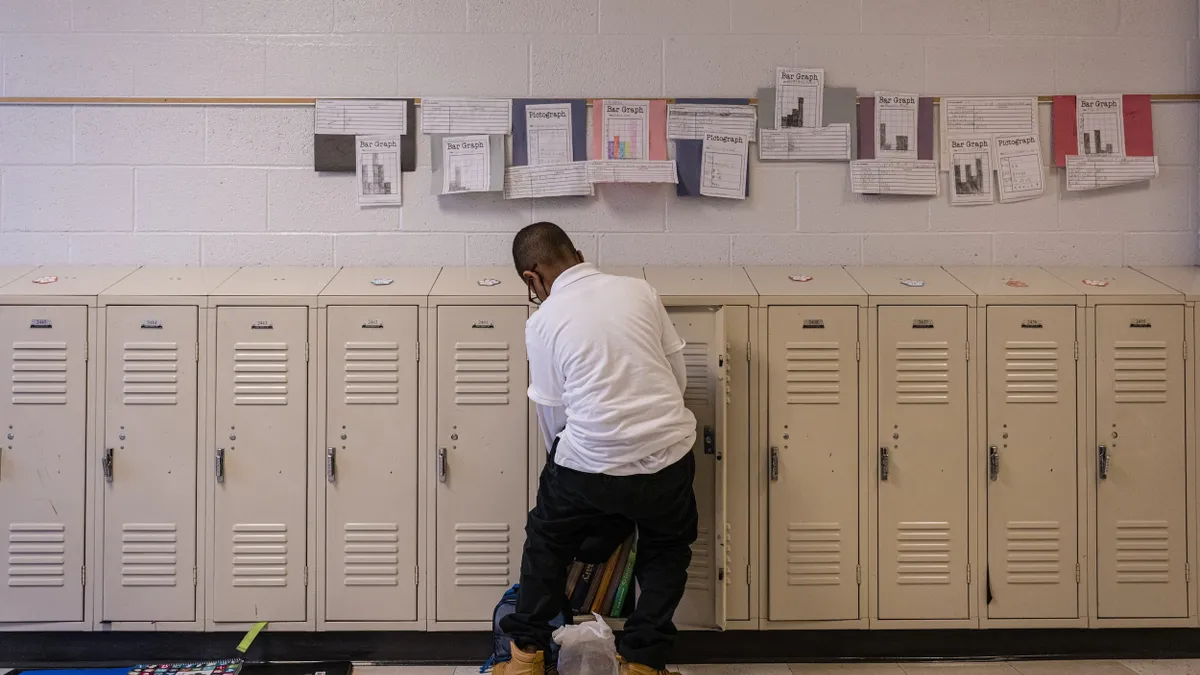 A student stands in front of a row of lockers with their back to the camera. The student is reaching into an open locker. There are materials like notebooks on the floor in front of the lockers.