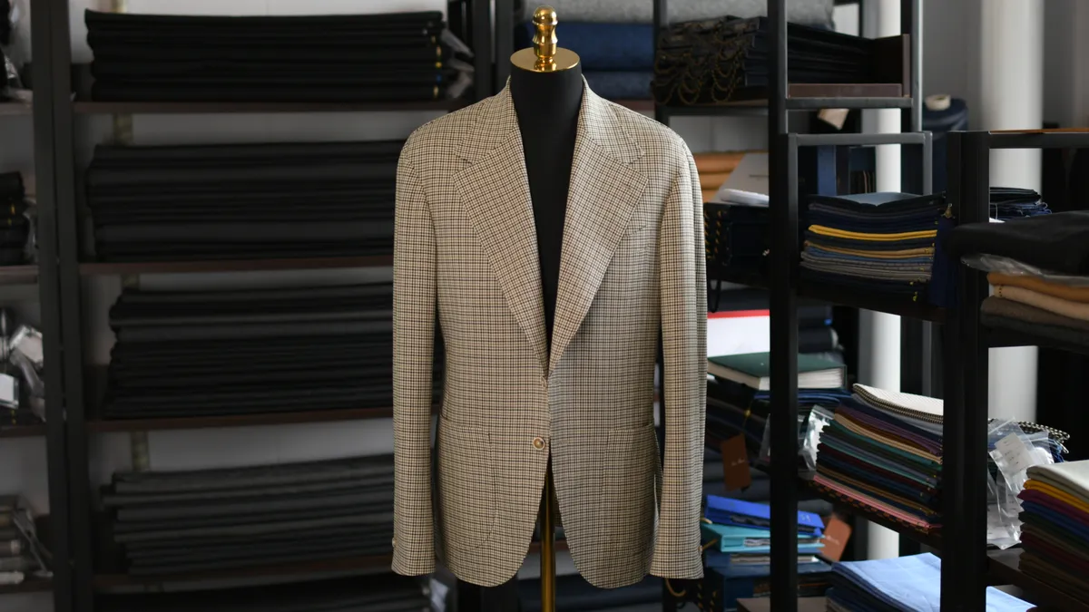 A brown tweed jacket hangs on a stand in a room full of wood shelves containing fabric samples.