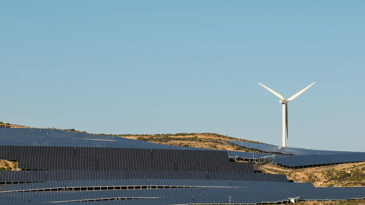 A large solar panel farm spread across a hillside with a single wind turbine in the background under a clear blue sky.