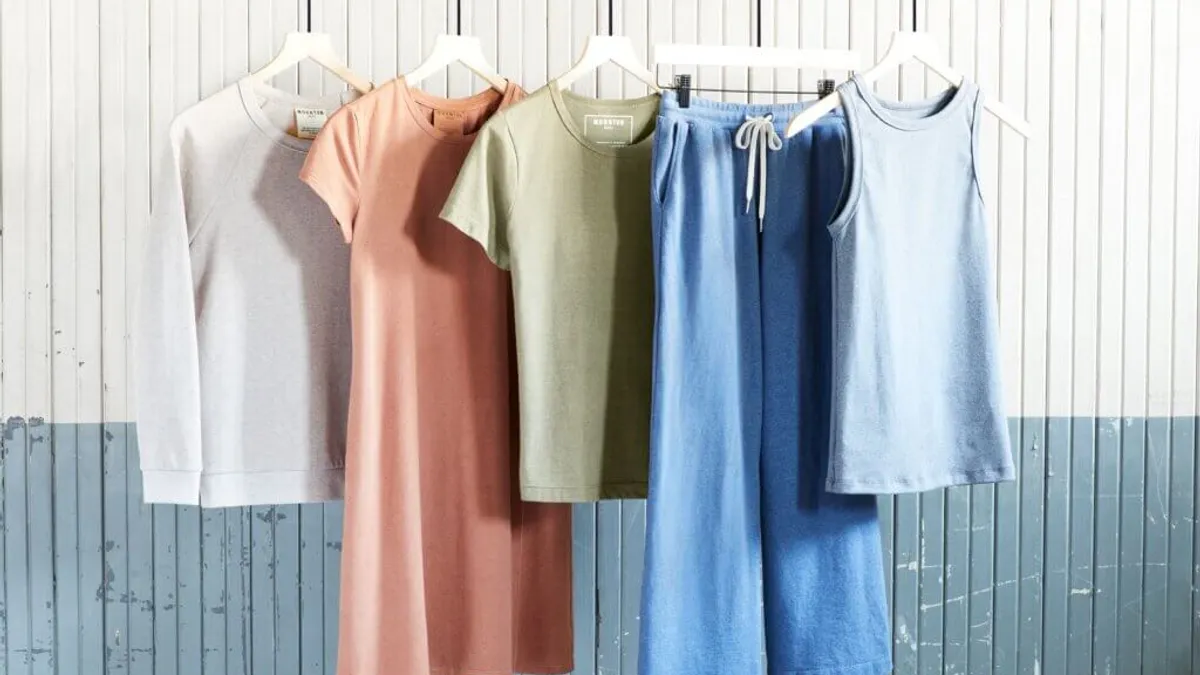 A white shirt, pink dress, green shirt, blue pants and blue tank on hangers, in front of a white and blue shiplap wall.