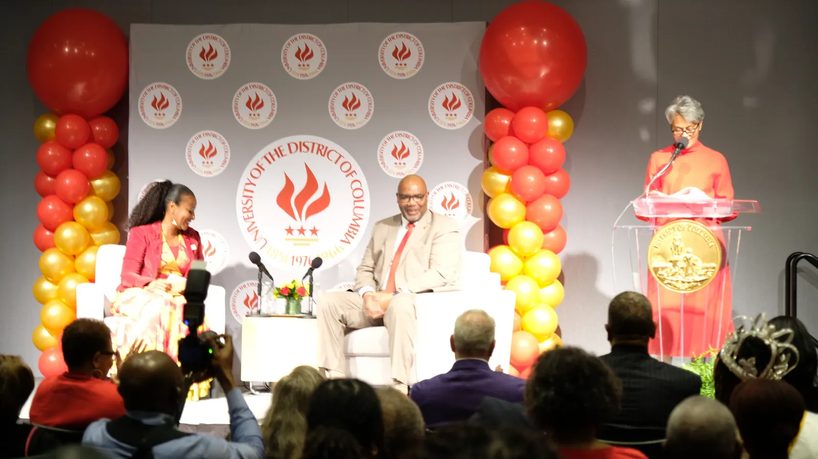 Man and woman sit across from each other on stage for fireside chat while another woman introduces from the podium.