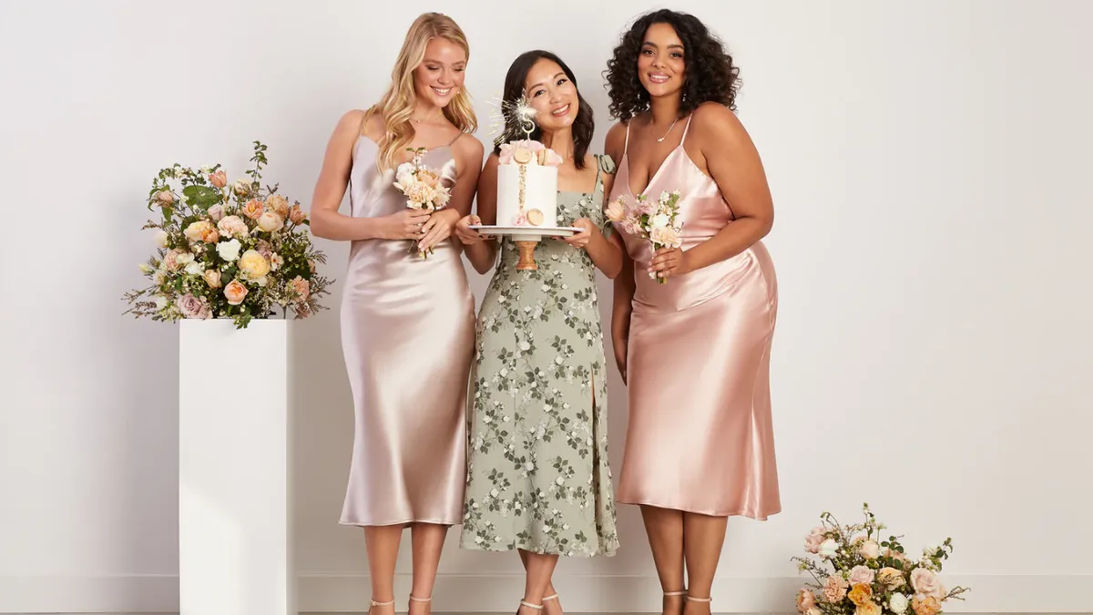 Three people wearing bridesmaid dresses stand together. The person in the center holds a cake.