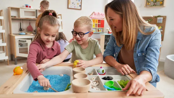 A teacher and two young students are working at an activity table in a classroom.
