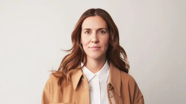 A person with long brown hair, white shirt and camel-colored jacket.