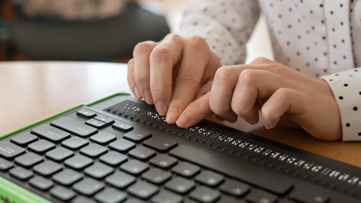 A person uses a computer with a Braille display and a computer keyboard. Inclusive device